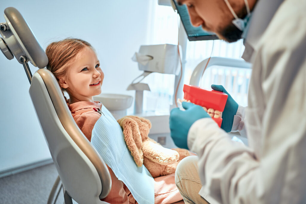 A little girl sits in a dental chair and looks at the doctor who shows her a model of the jaw and shows how to brush her teeth
